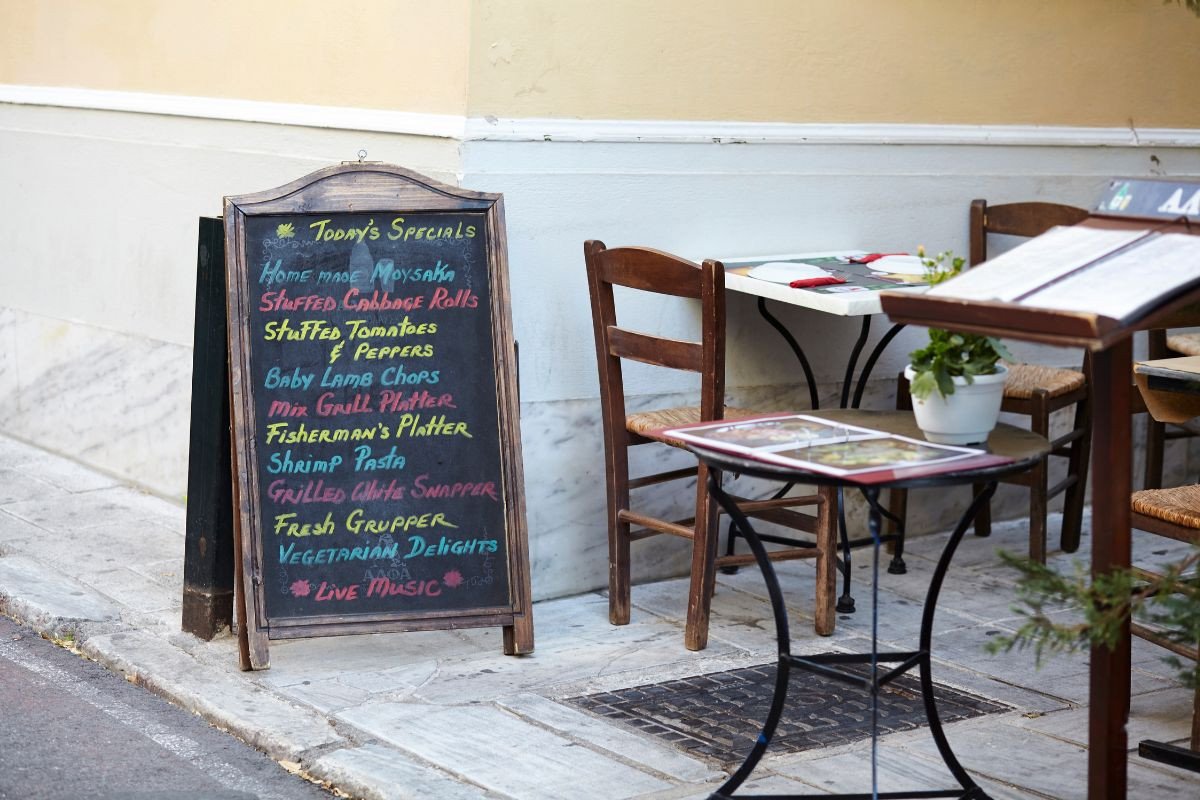A chalkboard outside a restaurant displays the day's specials in colorful chalk, including dishes like homemade moussaka, stuffed cabbage rolls, baby lamb chops, and grilled snapper. The board is positioned on a sidewalk next to a small table and chairs, creating an inviting atmosphere. The table has a menu and a small potted plant, while the restaurant’s wall is painted a soft yellow. The scene suggests a cozy and charming dining experience.